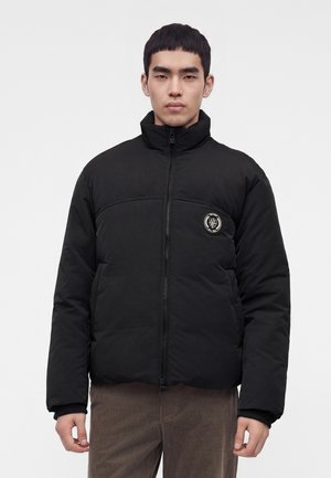 Homme portant une doudoune noire avec un col montant et un patch logo rond sur la poitrine, associé à un pantalon marron, debout devant un fond uni.