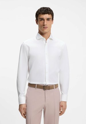 Ung mand iført en tætsiddende hvid skjorte, beige bukser og et brunt bælte, stående mod en ensfarvet hvid baggrund.