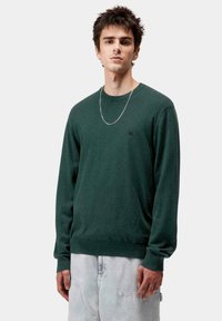 Jersey verde de cuello redondo hecho de una tela suave, con mangas largas y un dobladillo ajustado. Presenta un pequeño logo negro en el pecho.