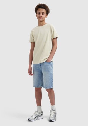 Teen boy standing with hand in pocket, wearing beige t-shirt, light denim shorts, white socks, and silver sneakers on plain background.
