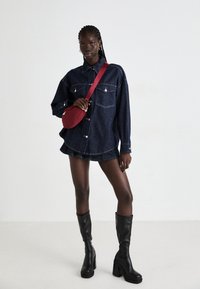 Abito camicia in denim con due tasche sul petto, colore blu scuro, abbinato a una borsa a tracolla rossa e stivali neri con tacco alto fino al ginocchio.
