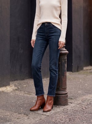 Personne portant un jean bleu foncé, un pull crème côtelé et des bottines marron, debout à l'extérieur près d'un poteau métallique usé dans une rue pavée.