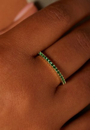 Goldener Ring mit grünen Edelsteinen, mit einem dünnen Band und einer Reihe abwechselnd kleiner und größerer Steine, der auf einer Hand ruht.