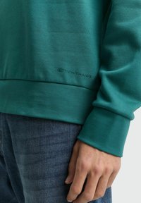 Felpa verde in cotone con maniche lunghe, caratterizzata da un polsino a coste e un piccolo logo impresso sull'orlo. Indossata con jeans blu.