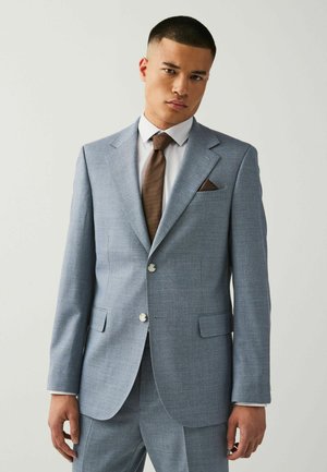 Hellblaues Sakko mit strukturierter Form, zwei Knöpfen und Revers. Mit einer braunen Krawatte und einem braunen Einstecktuch. Darunter ein elfenbeinfarbenes Hemd.