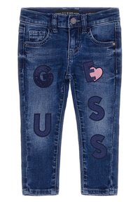 Vaqueros infantiles de color azul oscuro con letras "GUESS" y un parche de corazón roto rosa bordado en las patas delanteras.