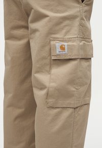 Μπεζ cargo παντελόνι Carhartt, εμφανιζόμενο από το μηρό μέχρι το γόνατο, με έμφαση σε μια μεγάλη τσέπη που φέρει το λογότυπο της μάρκας στη φάσα.