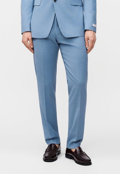 Άνδρας που φοράει ανοιχτό μπλε παντελόνι κοστουμιού και αντίστοιχο σακάκι με καφέ δερμάτινα μοκασίνια, στέκεται μπροστά σε απλό φόντο.