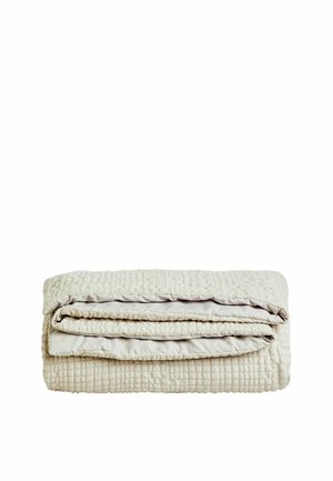 Couverture matelassée beige pliée avec un motif texturé en forme de grille sur fond blanc.