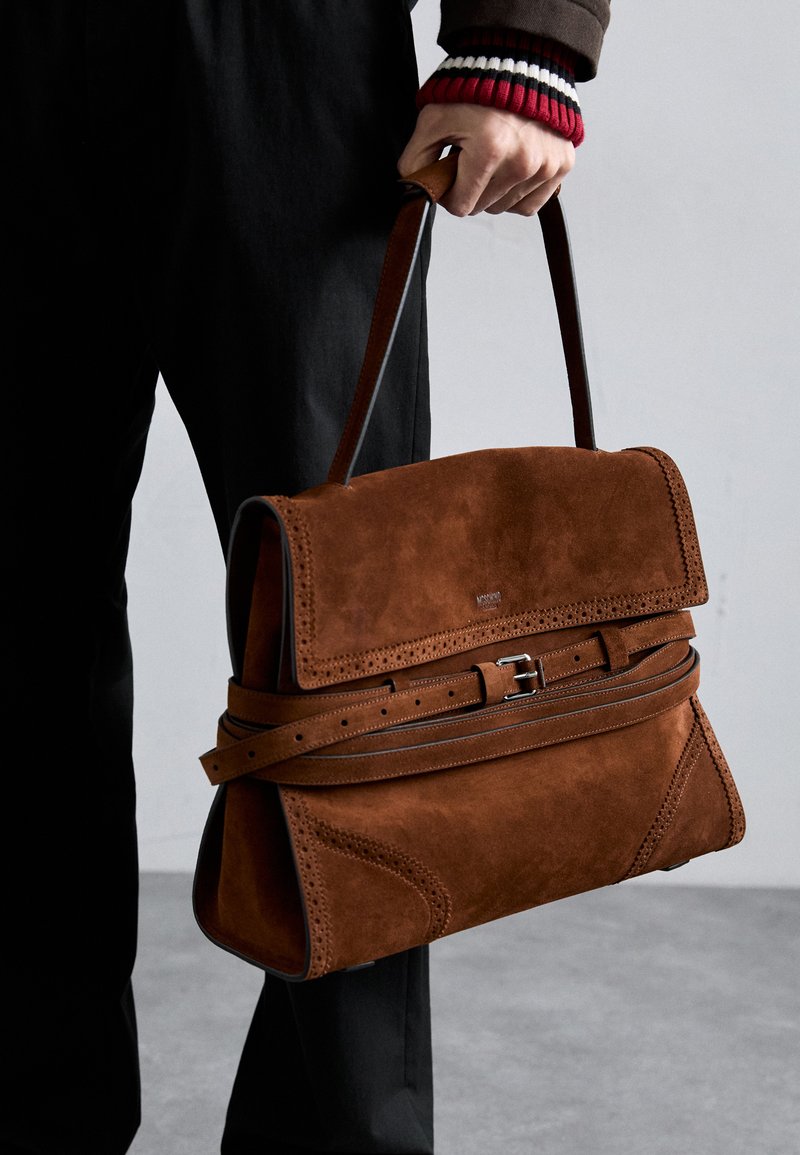 Brun mockaskinnie väska med vikdesign, utrustad med ett silverfärgat spänne och detaljerade perforeringar längs kanterna. Två robusta handtag.