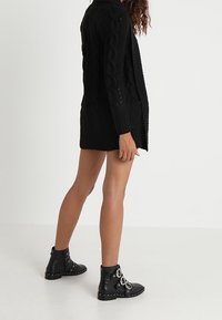 Czarny dziergany kardigan z wzorem warkoczowym, otwarty z przodu, z prążkowanym dołem i długimi rękawami. Noszony z czarnymi botkami do kostki z ozdobnymi ćwiekami.