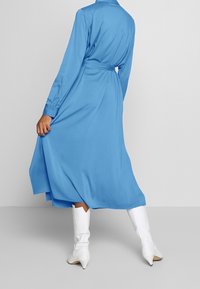 Robe à manches longues bleu clair avec col et taille ceinturée, silhouette fluide, associée à des bottes blanches à bouts pointus. Texture du tissu lisse.