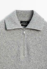 Maglione in lana grigia chiara a coste, con una cerniera corta e colletto ripiegato, che mostra un'etichetta del marchio all'interno del collo.
