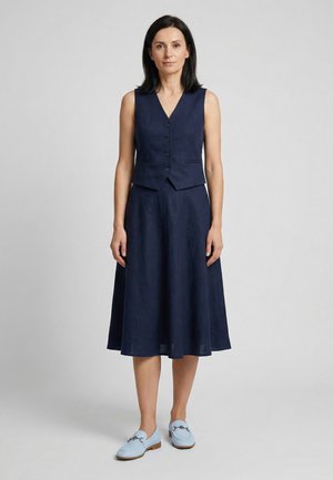 Vrouw staand, gekleed in een marineblauw mouwloos gilet met knopen en bijpassende midi-rok met lichtblauwe loafers op een effen achtergrond.