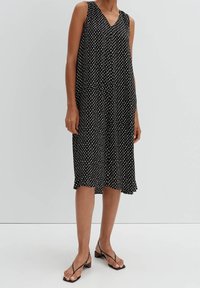 Mustapuku ilman hihoja, jossa on valkoisia pilkkuja, V-pääntie, virtaava siluetti, polven pituinen ja kevyt kangas. Minimalistinen muotoilu.