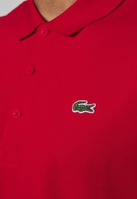 Red polo shirt with a textured fabric, featuring a classic collar, two buttons, and a green embroidered crocodile logo on the chest.