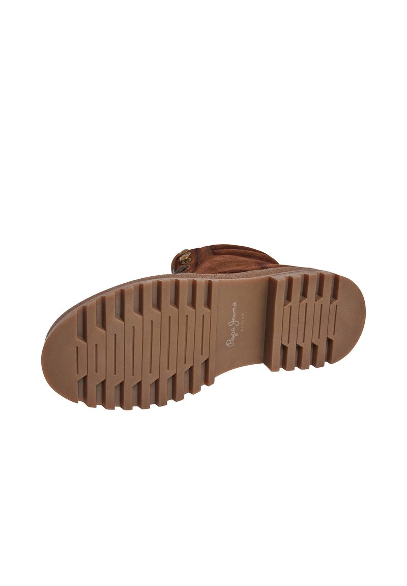 Bota de ante marrón con suela de goma texturizada, que presenta surcos horizontales y un logo. Acento de hardware dorado cerca de la parte superior.
