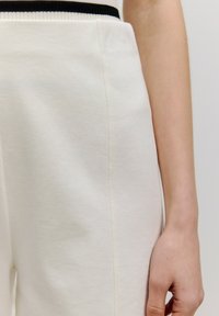 Weiße, strukturierte Stoffhosen mit einem schwarzen elastischen Bund. Nahaufnahme, die die Detailansicht der Seitennaht und einen Teil einer Hand hervorhebt.
