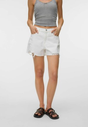 Woman wearing white frayed denim shorts, gray sleeveless top, and black sandals, standing with hands in pockets against a plain background.