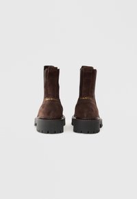 Botas de tobillo de ante marrón con paneles laterales elásticos, suelas de goma negra gruesas y un logotipo dorado en el tacón.