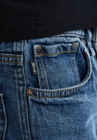 Jeans in denim blu con un primo piano sulla tasca frontale, caratterizzati da cuciture dorate e un piccolo etichetta del marchio. Tessuto liscio con una vestibilità classica.