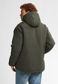 Olijfgroene pufferjack met capuchon, met een gladde, glanzende buitenkant en elastische manchetten, gecombineerd met lichtblauwe jeans.