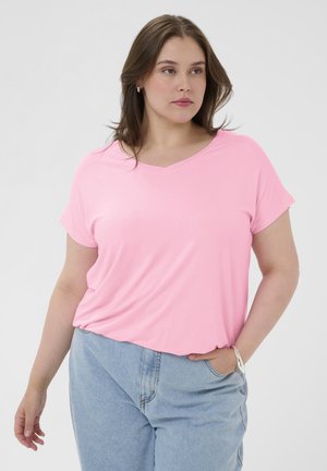 Donna con capelli castani a lunghezza spalle che indossa una T-shirt rosa chiaro con scollo a V e jeans azzurri, in piedi con una mano in tasca.