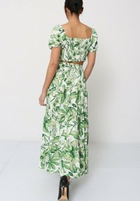 Robe en deux pièces avec des motifs floraux verts, des manches courtes bouffantes et une jupe maxi à volants avec un corsage ajusté. Portée avec des talons noirs.