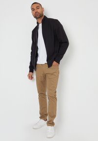 Zwarte ritssluitingsjack, wit T-shirt, tan broek en witte sneakers. Soepele stof, slim fit, minimalistisch ontwerp zonder versieringen of patronen.