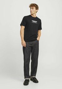 Črna bombažna majica s belim tiskom, v kombinaciji s temnimi kavbojkami in črnimi čevlji. Slim fit zasnova s kratkimi rokavi in krožnim izrezom.