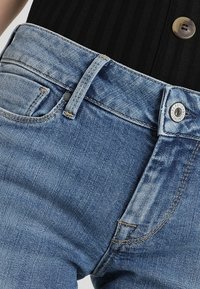 Vaqueros de mezclilla azul claro con una cintura de altura media, costuras visibles, cierre de botón y un pequeño detalle de bolsillo delantero. Tejido texturizado.