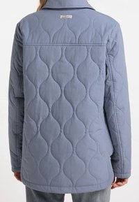 Pikowana kurtka w miękkim niebieskim materiale z wzorem fal, kołnierzem i metką na górnej części pleców. Długość sięga do bioder.