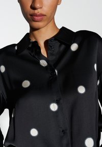 Zwarte blouse met grote witte stippen, knoopsluiting aan de voorkant en een kraag. Glad materiaal met een subtiele glans en een comfortabele pasvorm.