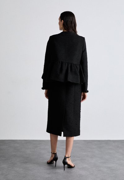 Black textured peplum jacket paired with a matching knee-length skirt, featuring a side slit. Worn with black high-heeled sandals.