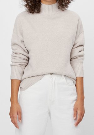 Personne portant un pull à col roulé côtelé beige clair avec les manches retroussées et un pantalon blanc taille haute sur un fond neutre.