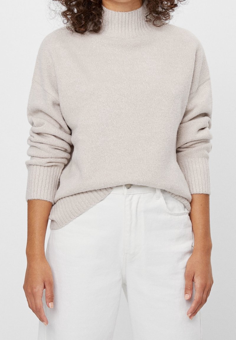 Personne portant un pull à col roulé côtelé beige clair avec les manches retroussées et un pantalon blanc taille haute sur un fond neutre.