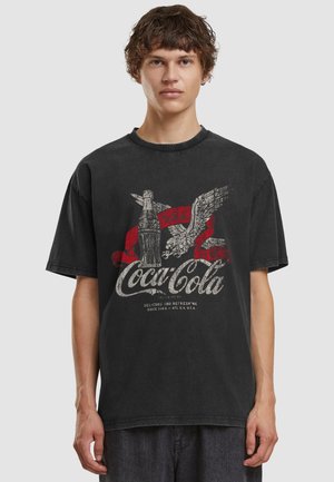 Junger Mann trägt ein ausgewaschenes schwarzes Coca-Cola T-Shirt mit Vintage-Flasche und Adler-Grafik, steht vor schlichtem Hintergrund.