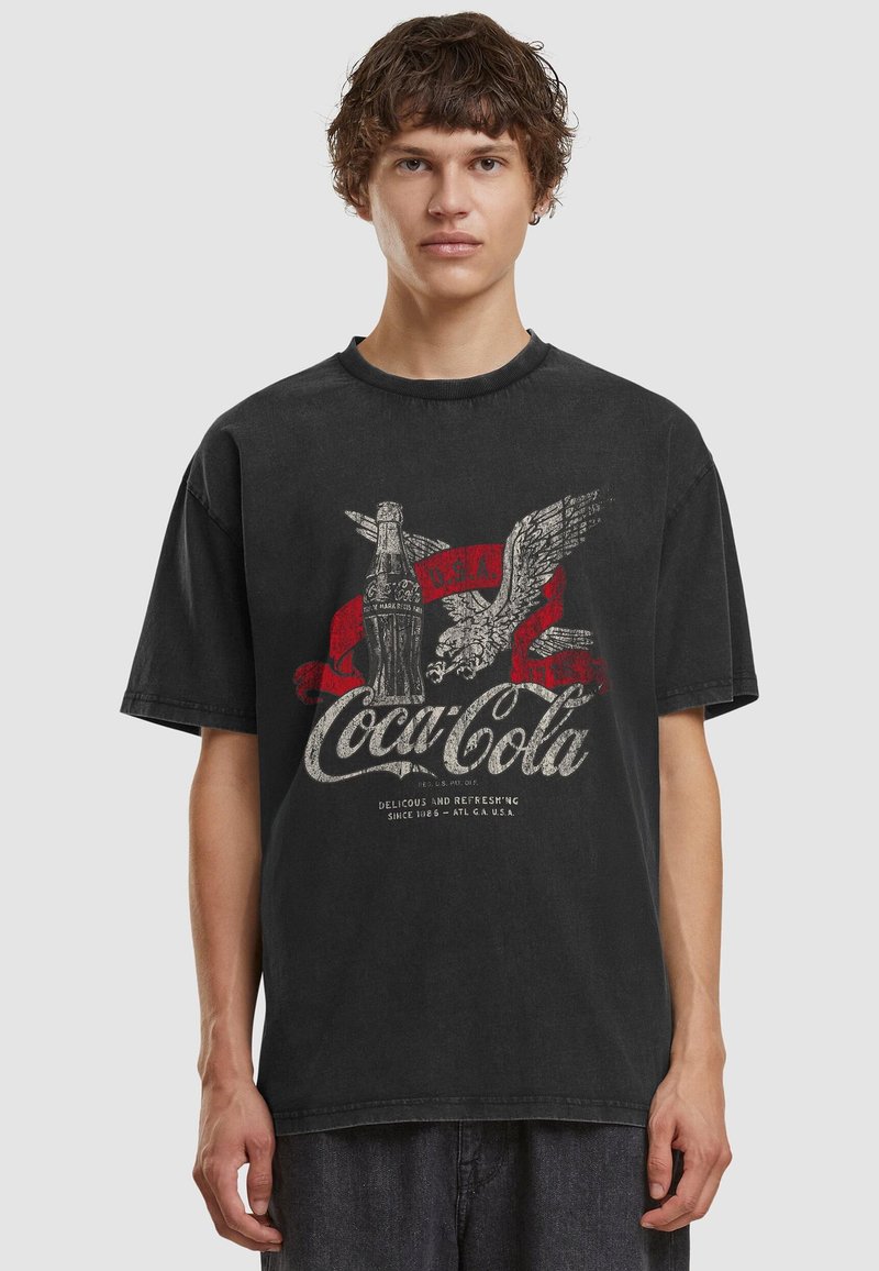 Νέος άνδρας φοράει ξεθωριασμένο μαύρο t-shirt Coca-Cola με γραφικό από vintage μπουκάλι και αετό, στέκεται μπροστά σε απλό φόντο.