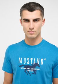 Turkusowa bawełniana koszulka z krótkim rękawem, z białym logo "MUSTANG" i grafiką konia na środku klatki piersiowej.