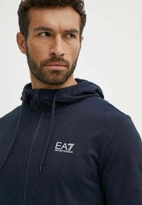 Felpa con zip blu navy e cappuccio regolabile, con logo EA7 bianco sul lato sinistro del petto, realizzata in un tessuto morbido e con una texture liscia.