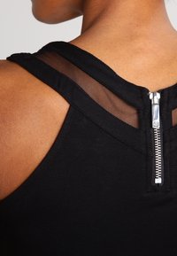 Sort ærmeløs top med gennemsigtige meshdetaljer på skuldrene og en sølvfarvet lynlås bagpå. Glat, tætsiddende tekstur.