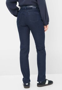 Jeans slim fit blu scuro con una superficie liscia, dotati di due tasche posteriori e una cintura, abbinati a sneaker nere e a una maglietta navy.