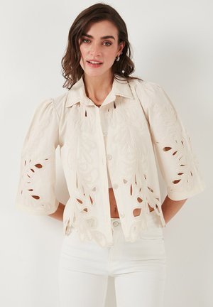 Cream-colored blouse with openwork leaf designs, short puff sleeves, and a button-down front, made from lightweight fabric.