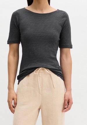 Femme portant un haut gris foncé près du corps à manches courtes et un pantalon ample beige clair avec une taille à cordon noué, sur un fond uni.
