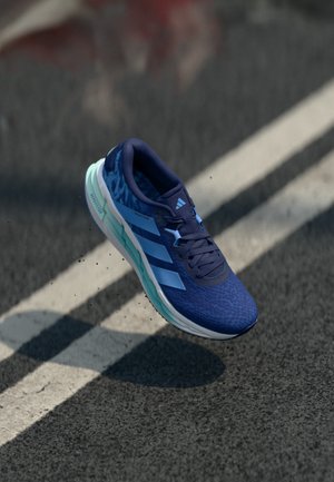 Blue athletic shoe with a textured mesh upper, light blue accents, and a cushioned sole. Design features three white stripes on the side.