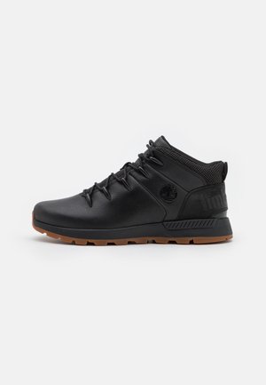 Botte mi-haute en cuir noir avec lacets noirs, col rembourré à la cheville et semelle en caoutchouc marron, vue de côté extérieur sur fond blanc.