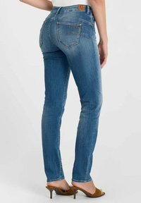 Strakke blauwe spijkerbroek van denim, met een achterontwerp met twee zakken en gestikte details. Draagt bruine sandalen met hak.