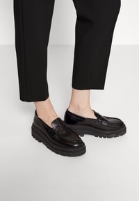 Mocasines de cuero negro con una suela plataforma gruesa, con un acabado liso y detalles de costura tradicionales. Usados con pantalones negros recortados.