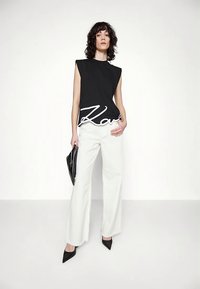 Žena s kudrnatými vlasy, oblečená v černém topu bez rukávů s bílým nápisem, širokých bílých kalhotách, černých špičatých podpatcích a držící černou kabelku.