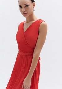 Rode mouwloze jurk met geplisseerde stof, v-hals en een getailleerde taille. Heeft een ceintuur in de taille en een vloeiende rok. Gladde textuur.